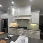 Modern kitchen featuring white cabinetry, stainless steel appliances, and a large island with a dark countertop, illuminated by pendant lighting, showcasing custom cabinetry and remodeling services by The Perfect Cabinet Company.