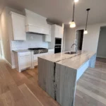 Modern kitchen interior featuring a large island with a wooden countertop, white cabinetry, stainless steel appliances, and pendant lighting, showcasing The Perfect Cabinet Company's custom cabinetry and remodeling services.