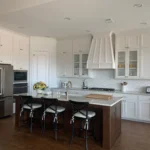 Modern kitchen with white cabinetry, dark island, stainless steel appliances, and bar stools, showcasing The Perfect Cabinet's remodeling and custom cabinetry services.