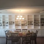 Elegant dining area featuring a round table surrounded by wooden chairs, complemented by a bright chandelier and white cabinetry with shelves displaying decorative items, showcasing The Perfect Cabinet's craftsmanship in home remodeling.