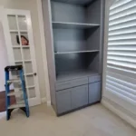 Modern gray cabinetry with open shelves and drawers in a well-lit office space, featuring a ladder and window with blinds.