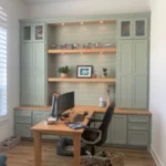Office setup featuring custom cabinetry in soft green, wooden desk with computer, and decorative shelves, showcasing The Perfect Cabinet's design capabilities.