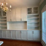 Office cabinetry design featuring white shelves and cabinets, elegant chandelier, and natural light from window, showcasing The Perfect Cabinet's remodeling expertise.