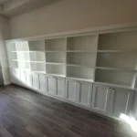 White cabinetry with open shelves and closed storage at the bottom, set against a neutral wall in an office space.