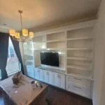Modern office cabinetry with built-in shelves and TV, featuring a chandelier and a wooden table, showcasing The Perfect Cabinet's design expertise.