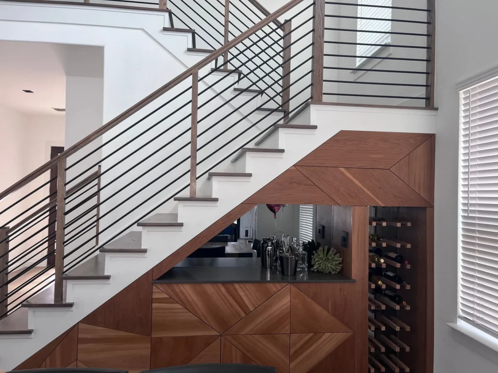 Modern staircase with wooden handrails, sleek metal balusters, and a custom cabinetry bar area featuring geometric wood design and a wine rack.