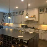 Custom kitchen island with dark countertop and wine storage, surrounded by white cabinetry and pendant lighting, showcasing a modern kitchen design by The Perfect Cabinet.
