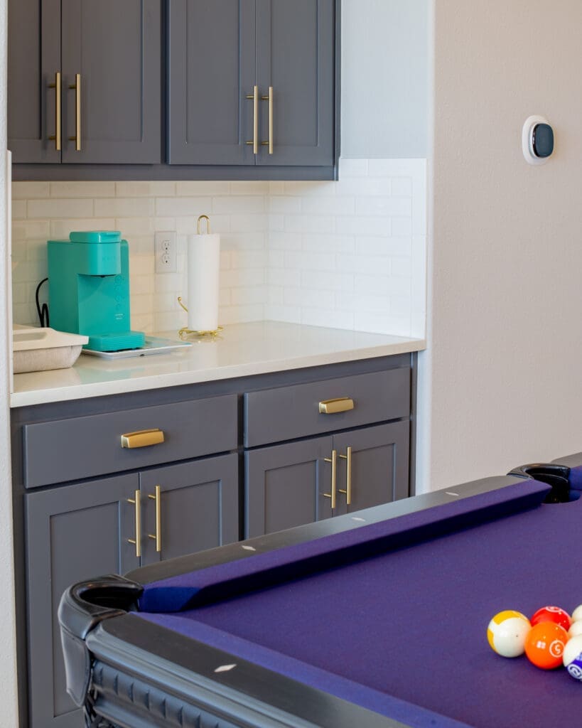 Modern kitchen cabinetry with gray doors and gold handles, featuring a turquoise coffee maker and a white paper towel holder on a countertop, with a pool table in the foreground showcasing colorful billiard balls.