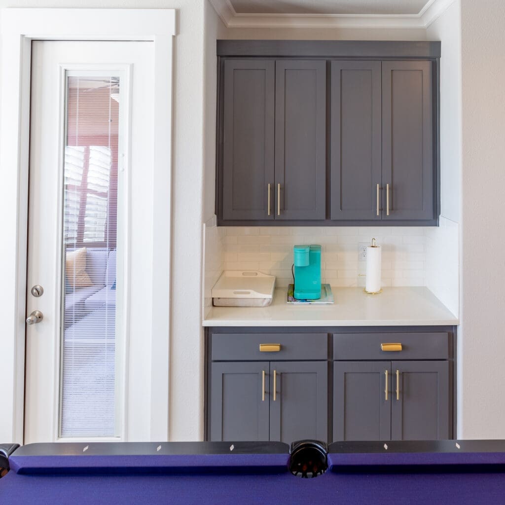 Modern gray cabinetry with gold hardware, countertop with turquoise pitcher and paper towel holder, adjacent to a glass door in a home remodeling setting.
