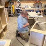 Man kneeling in workshop assembling custom cabinetry with tools and materials visible.