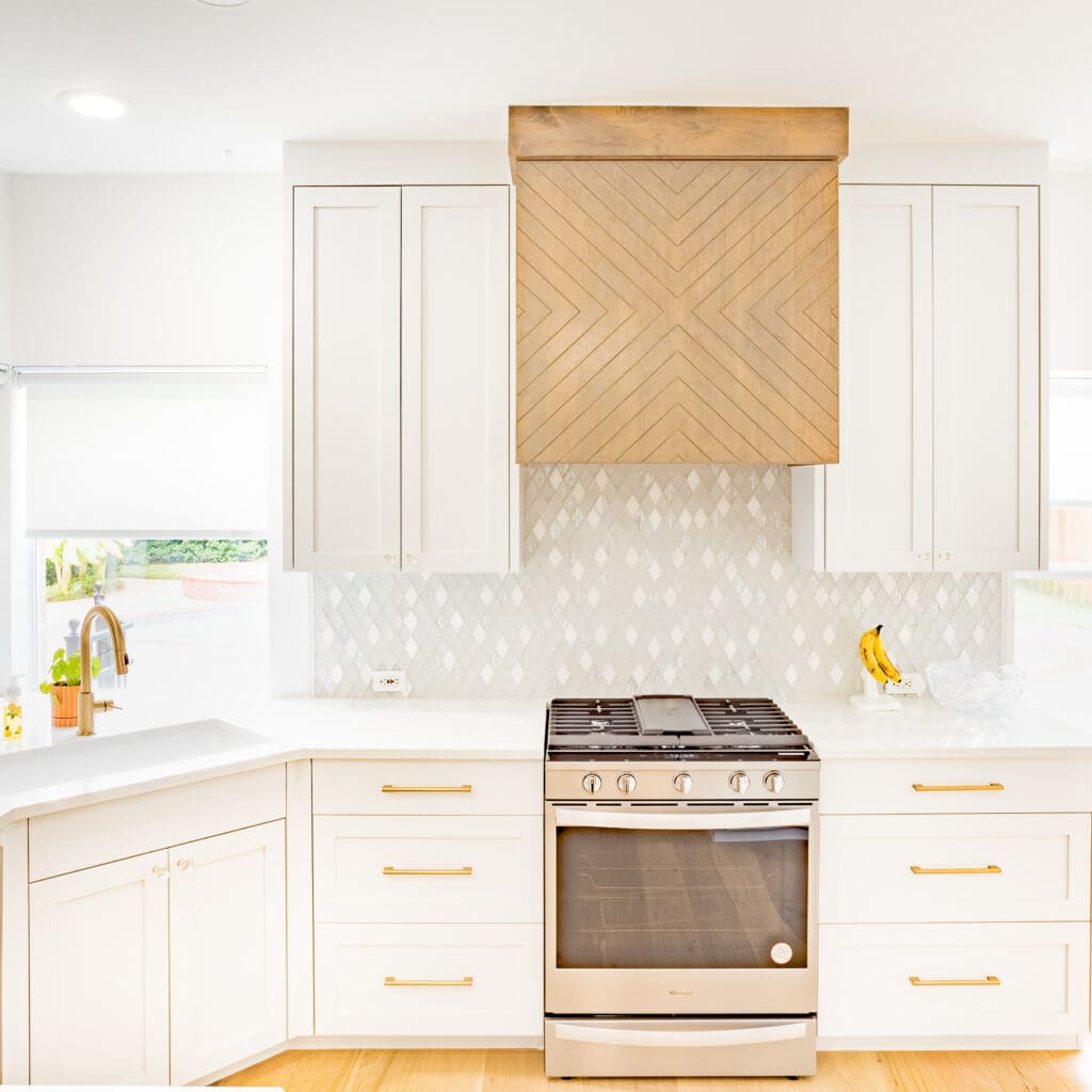Modern kitchen featuring white cabinetry, a wood-patterned range hood, stainless steel stove, and light marble backsplash, ideal for home remodeling inspiration.