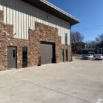 Exterior view of The Perfect Cabinet Shop building featuring a stone facade, large garage door, and clear blue sky, showcasing the business's custom cabinetry services.