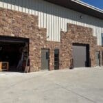 Exterior view of The Perfect Cabinet Shop showcasing stone and metal architecture, garage doors, and a clear sky, emphasizing custom cabinetry services.
