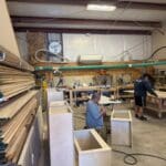 Workshop interior showcasing custom cabinetry production, featuring workers assembling cabinets and wooden materials organized on shelves.