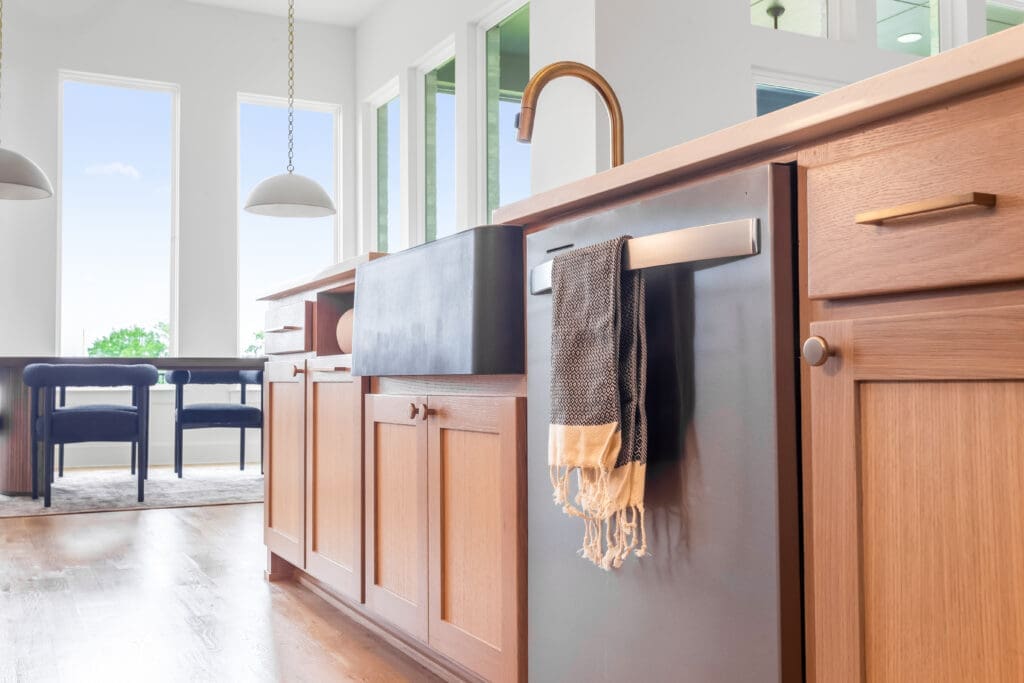 Modern kitchen featuring custom cabinetry, a stylish sink, and a dish towel hanging on the dishwasher, with large windows allowing natural light and a view of outdoor greenery.