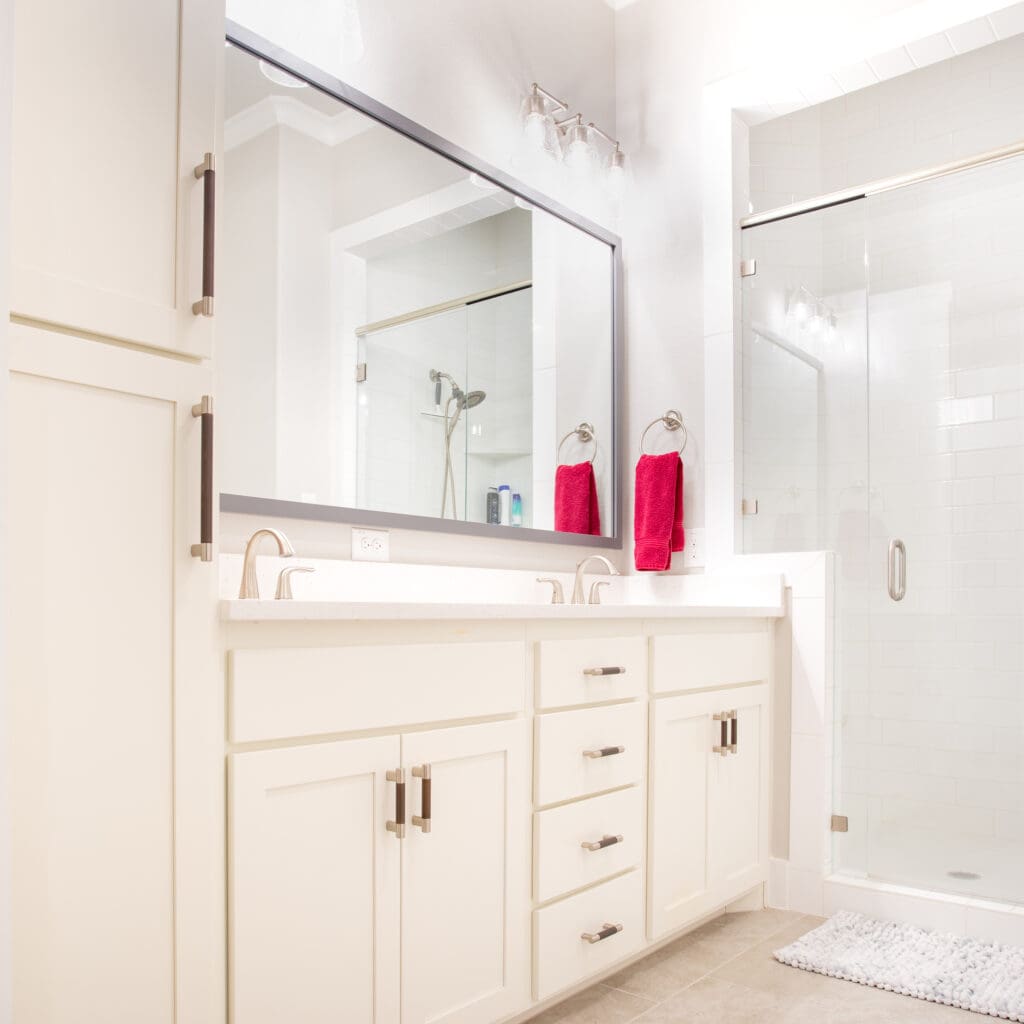 Modern bathroom featuring white cabinetry, large mirror, dual sinks, and glass shower enclosure, with pink towels and neutral decor, showcasing home remodeling design by The Perfect Cabinet Company.