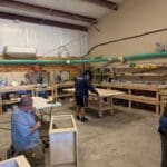 Cabinet makers working in a woodworking shop, assembling custom cabinetry amidst tools and materials, showcasing craftsmanship relevant to The Perfect Cabinet Shop.