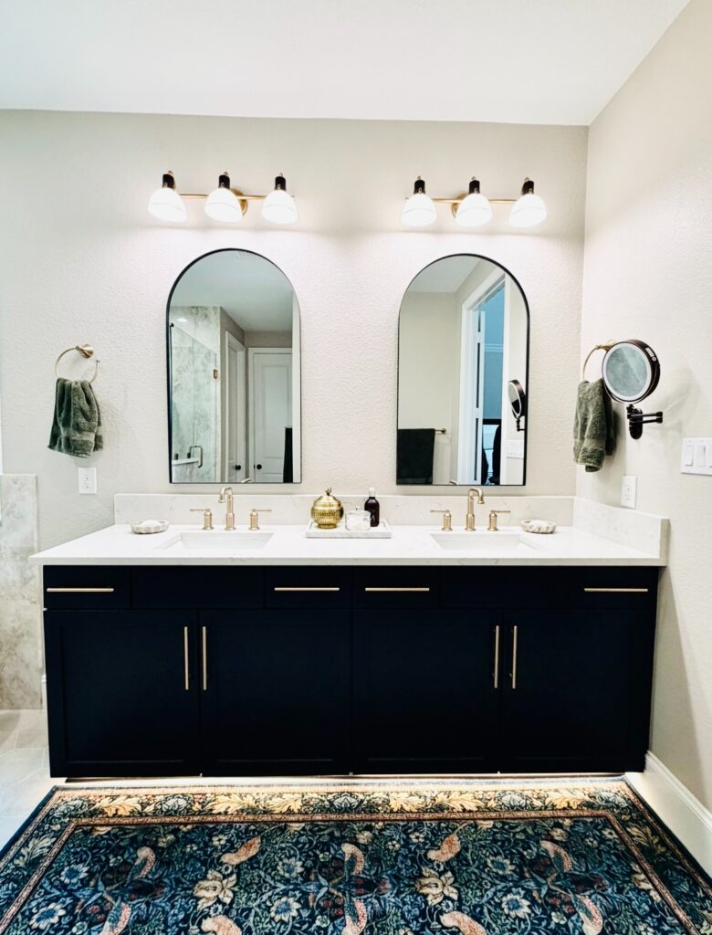 Modern bathroom vanity with dual sinks, gold fixtures, arched mirrors, and decorative rug, showcasing custom cabinetry and contemporary design elements.