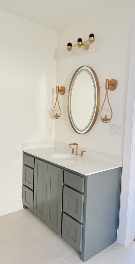 Modern bathroom vanity with gray cabinetry, white countertop, round mirror, and stylish copper lighting fixtures.