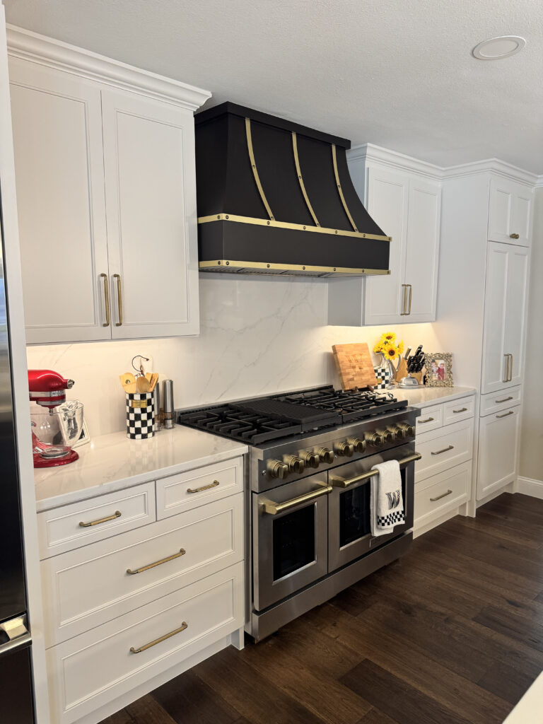 Modern kitchen featuring coastal theme cabinetry, a large gas range with a black and gold hood, white cabinets, and a marble backsplash, ideal for kitchen remodeling inspiration in Collin County and DFW.