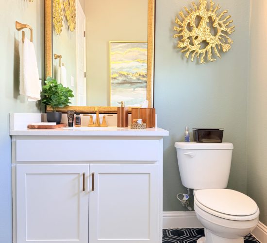 Modern open concept bathroom featuring white cabinetry, gold accents, and a decorative mirror, with a green wall, stylish artwork, and organized countertop storage.