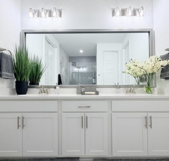 Modern open concept bathroom with dual sinks, white cabinetry, decorative plants, and frameless mirror reflecting a spacious design, emphasizing functionality and spa-like aesthetics.