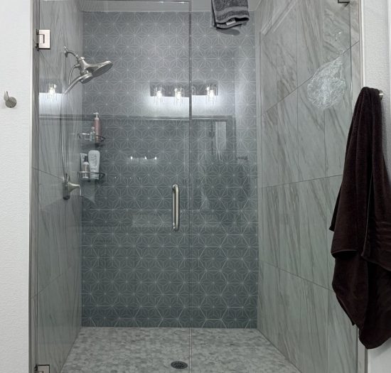 Modern glass shower enclosure with geometric tile backdrop, featuring chrome showerhead and wall-mounted storage for toiletries, complemented by a plush dark towel hanging nearby.