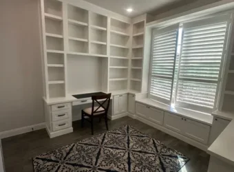 Modern home office with custom white cabinetry, built-in shelves, and a desk, featuring a patterned rug and window providing natural light.
